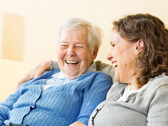 image of senior woman and mature woman/caregivier, sitting on sofa and laughing together